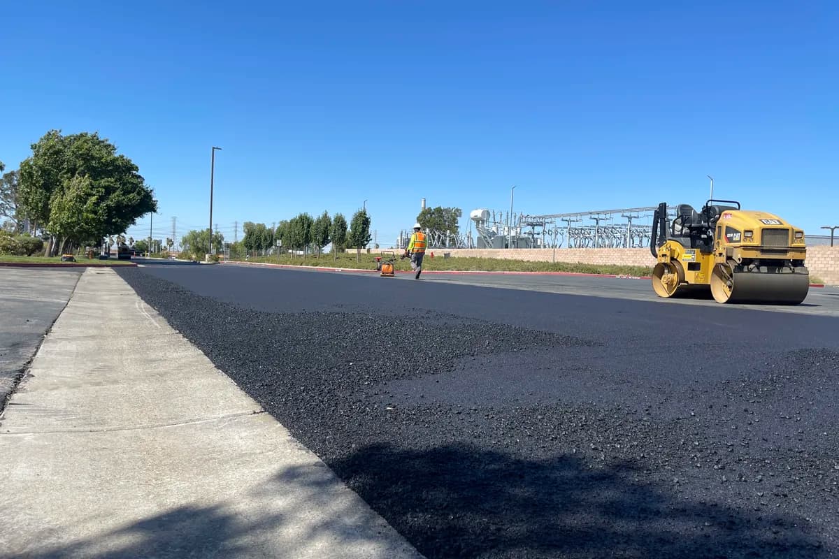 Roller compacting a freshly placed asphalt mat next to a concrete walkway on a commercial paving project.