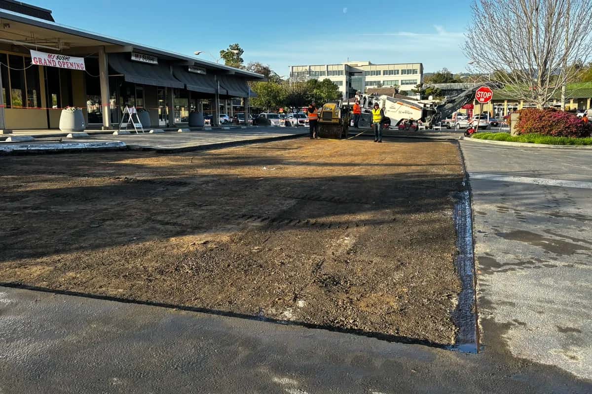 Milled-out section of a commercial parking lot prepped for an asphalt patch, with a cold planer and roller working the repair area.