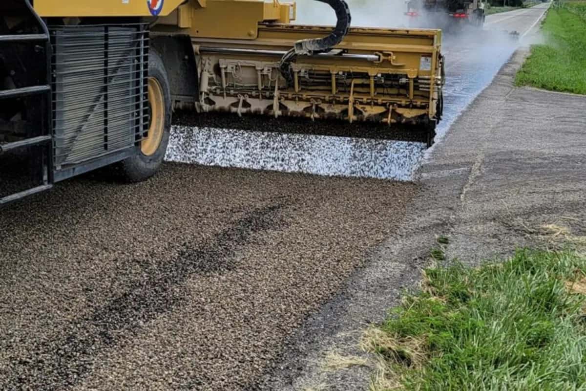 Chip seal spreader box laying aggregate over fresh asphalt emulsion on a rural road, with a distributor truck applying binder ahead.