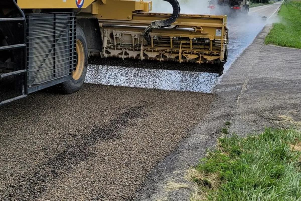 Chip seal spreader box laying aggregate over fresh asphalt emulsion on a rural road, with a distributor truck applying binder ahead.