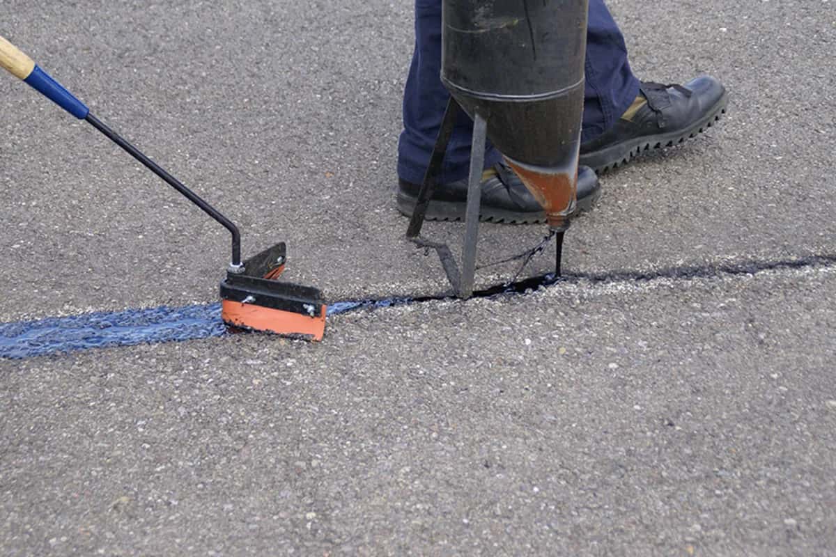 Close-up of a worker applying hot rubberized crack sealant into a pavement crack with a wand and squeegee applicator.