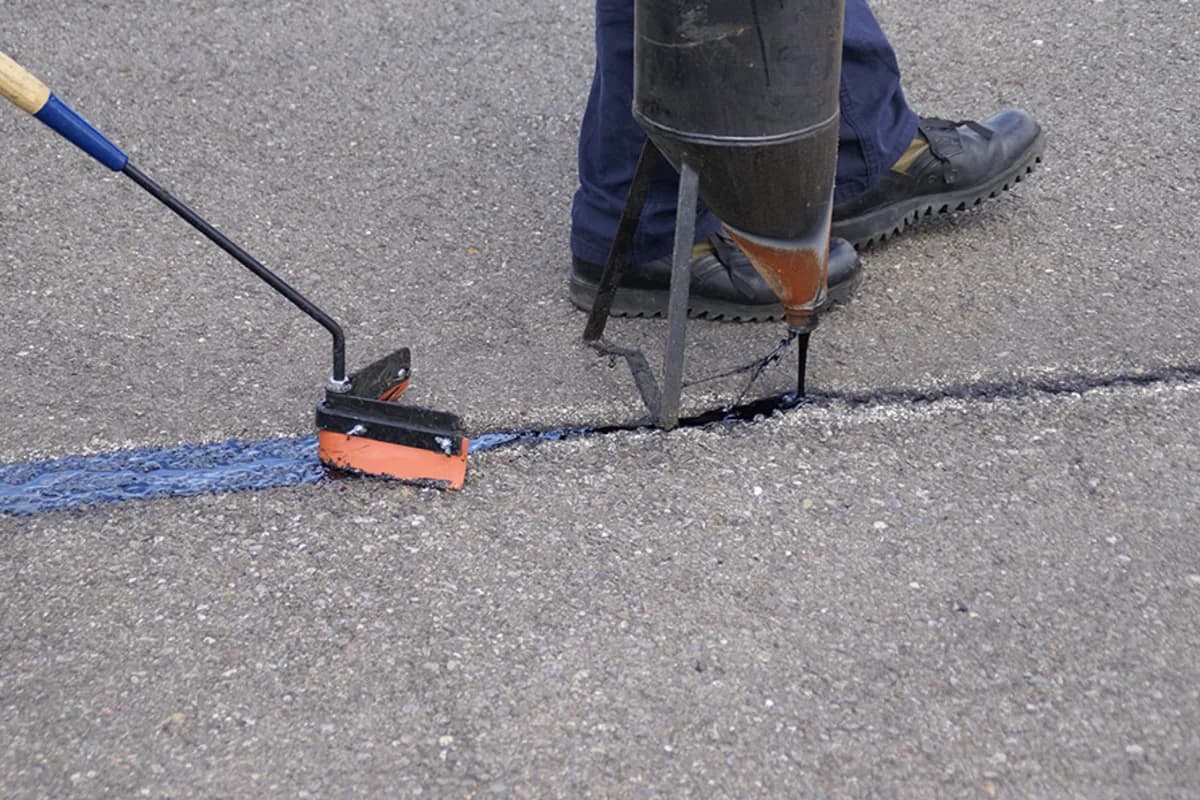 Close-up of a worker applying hot rubberized crack sealant into a pavement crack with a wand and squeegee applicator.