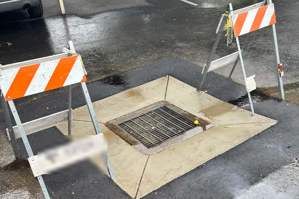 A parking lot catch basin and drain inlet reset flush with freshly placed asphalt, marked off with traffic barricades.