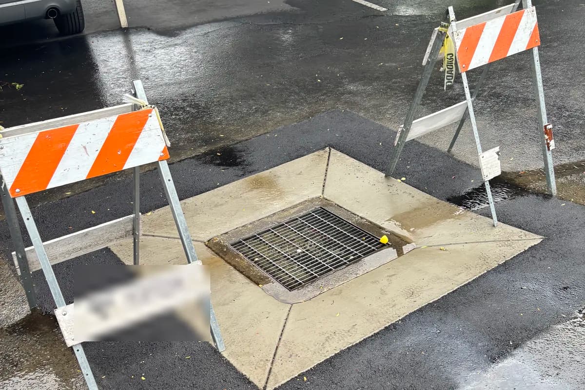 A parking lot catch basin and drain inlet reset flush with freshly placed asphalt, marked off with traffic barricades.