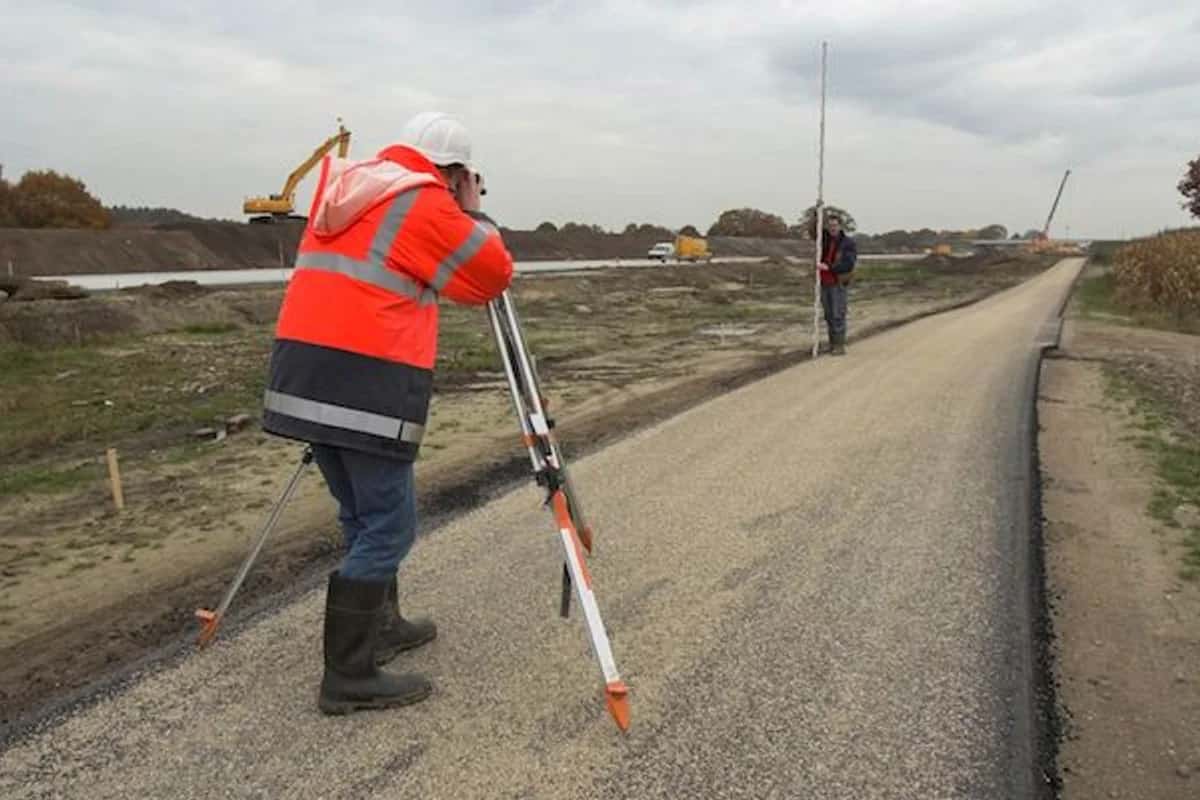 Surveyor using a level and tripod to check grade on a freshly paved road under construction, with a second worker holding a grade rod down the road.