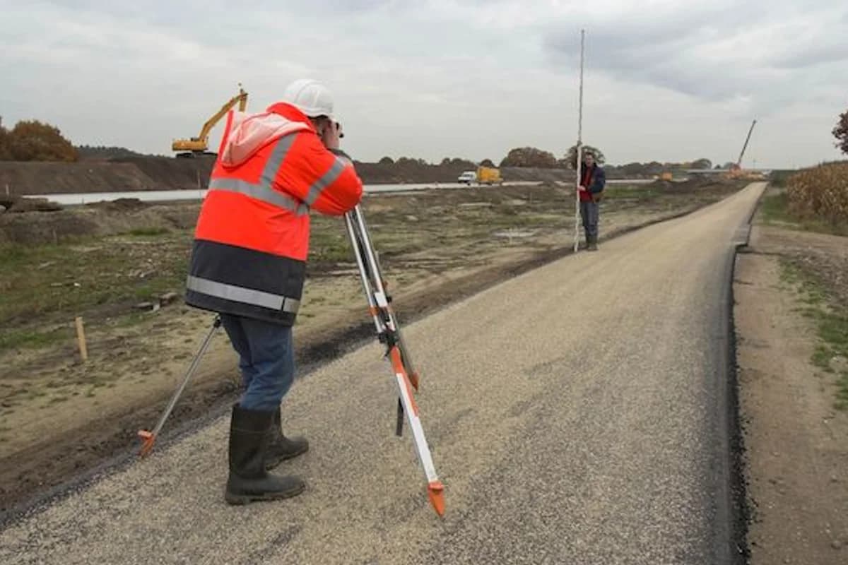 Surveyor using a level and tripod to check grade on a freshly paved road under construction, with a second worker holding a grade rod down the road.