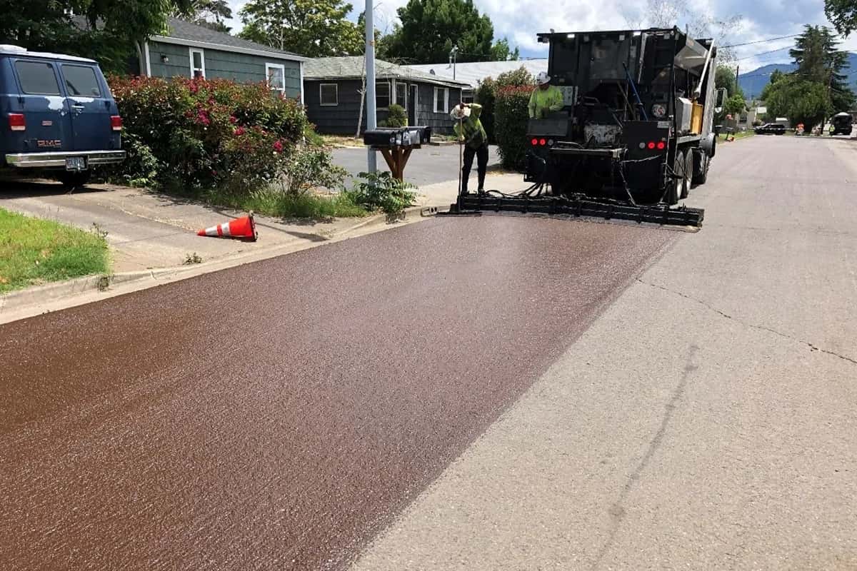 Spreader box laying a fresh slurry seal across a residential street, the new surface treatment meeting the untreated asphalt along a clean straight edge.