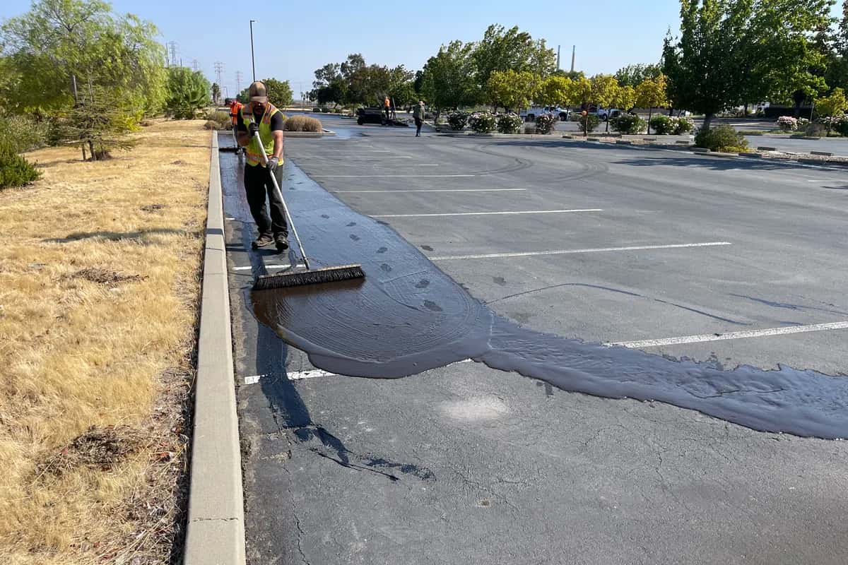 Worker applying sealcoat with a squeegee, leaving a fresh dark coat next to faded asphalt in a commercial lot.