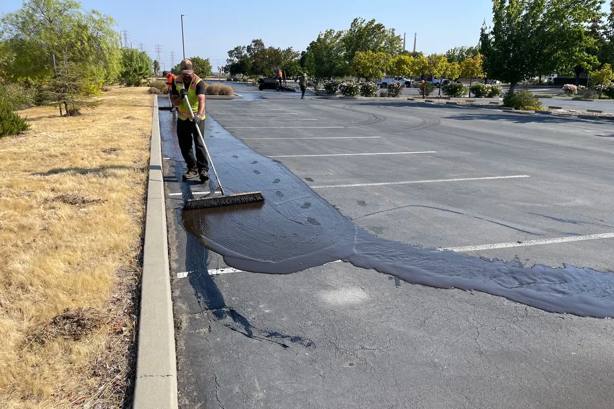 Worker applying sealcoat with a squeegee, leaving a fresh dark coat next to faded asphalt in a commercial lot.