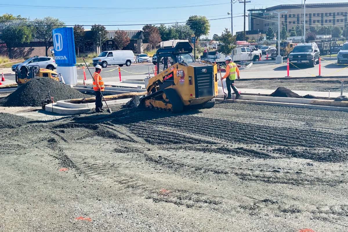 Track loader spreading and grading aggregate base across a commercial lot subgrade, with a crew raking the edges and a roller standing by ahead of paving.