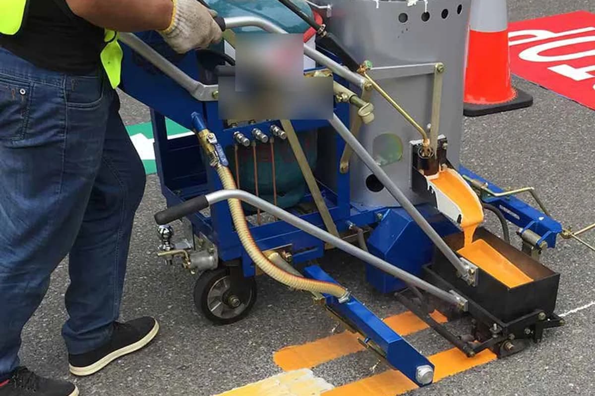 Worker operating a walk-behind thermoplastic striping machine, applying a fresh yellow thermoplastic line to a road surface.