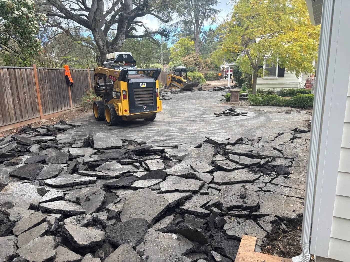 Broken asphalt lifted from a residential driveway during full-depth removal, with a skid steer clearing debris