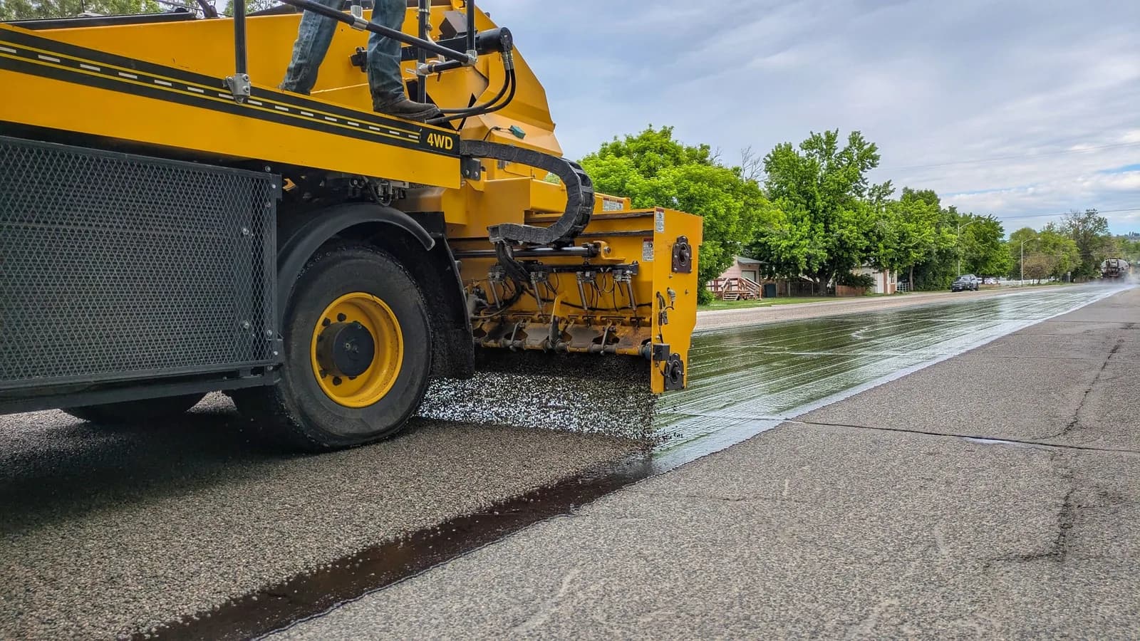 A chip spreader discharging aggregate onto fresh binder, with the newly chipped surface behind it meeting the older pavement it is being placed over.