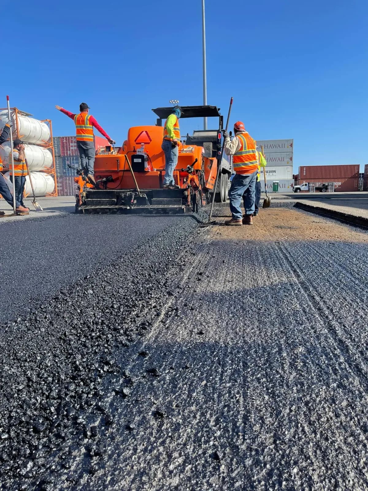 A paving crew placing and raking hot mix asphalt across an industrial yard behind a paver