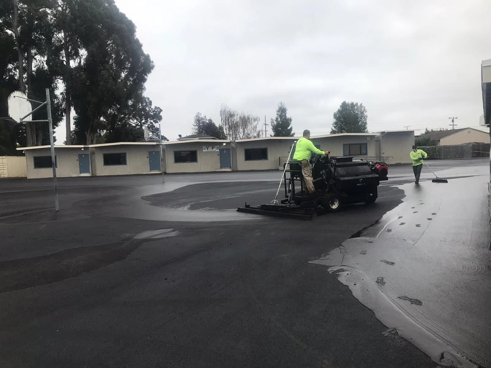 A crew applying sealcoat across a parking lot with a squeegee machine while a second worker cuts in by hand