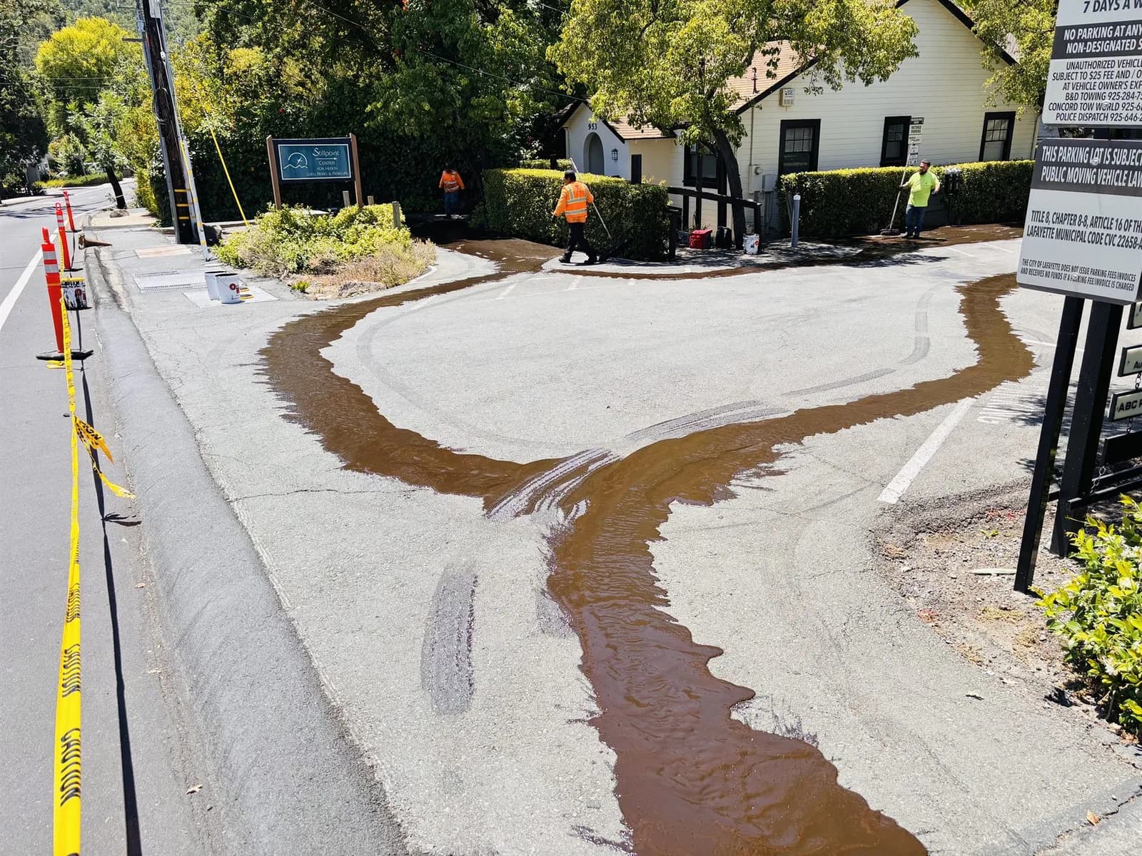 Brown sealer flowing across the edge of a parking lot as a crew works it in by hand near the street