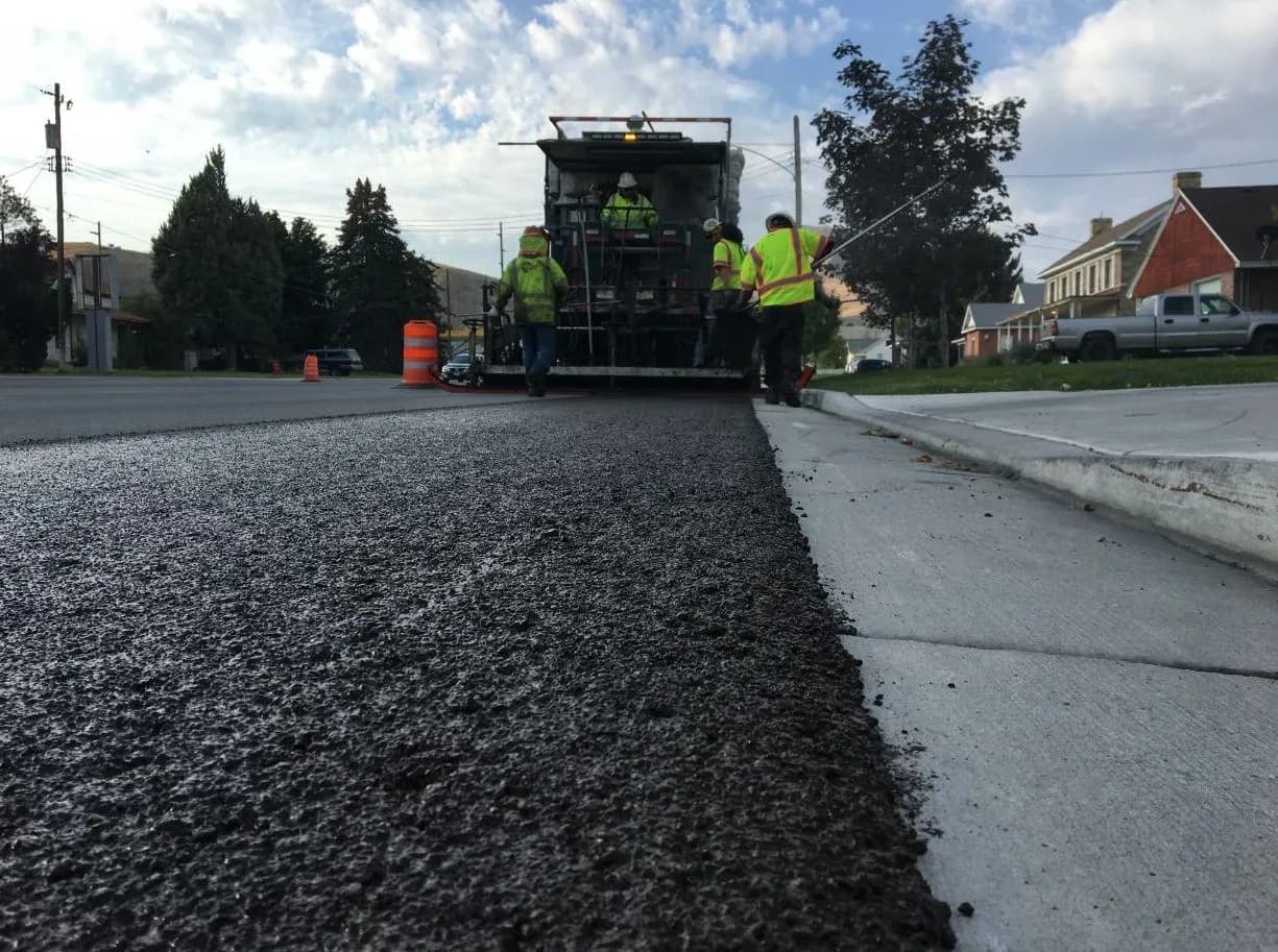 Ground-level view along a freshly placed slurry seal mat, its coarse aggregate texture meeting the concrete gutter, with the spreader machine and crew working ahead.