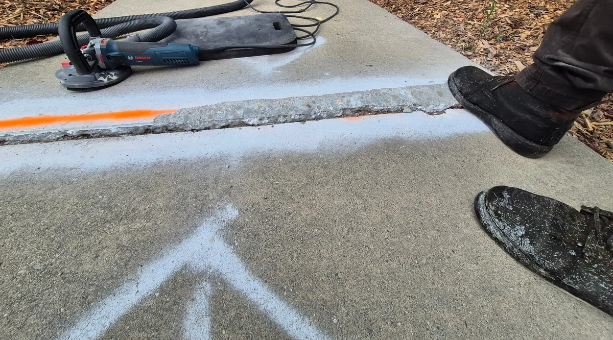 A sidewalk panel offset roughly an inch above the adjoining slab, with the raised edge outlined in orange and white marking paint, a pedestrian's boot at the lip, and a dust-shrouded concrete grinder staged on the high slab ready to cut the edge down.